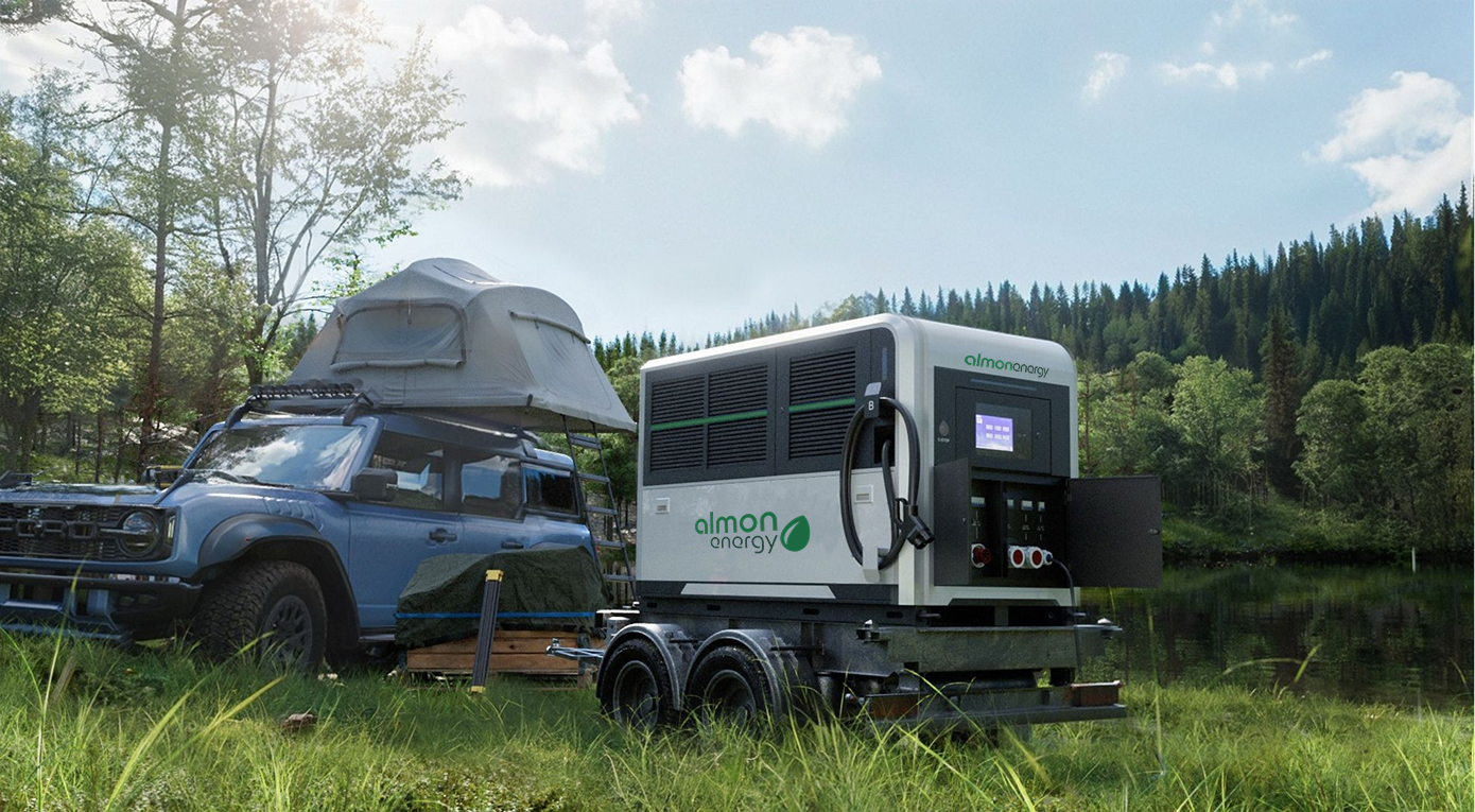 Ein blauer, geländegängiger SUV mit Dachzelt steht neben einer auf einem Anhänger montierten mobilen Ladestation von Almon Energy. Die Szene spielt in einer natürlichen Umgebung mit hohem Gras, einem See oder Fluss und einem dichten Wald unter hellem, sonnigem Himmel.
