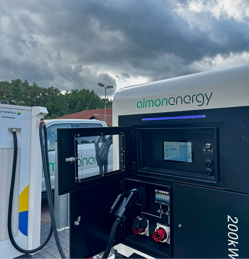 Geöffnete Serviceklappe des mobilen 200-kWh-Batteriespeichers von Almon Energy mit Display, CEE-Steckdosen und CCS-Ladekabel neben einer Vattenfall-Ladesäule.