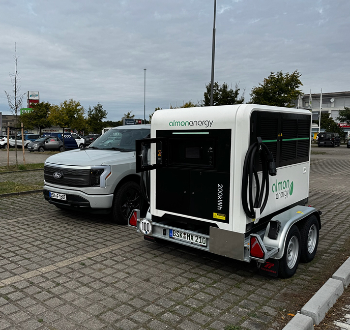 Mobiler 200-kWh-Batterie-Energiespeicher von Almon Energy auf einem PKW-Anhänger, geparkt neben einem elektrischen Ford F-150 Lightning Pickup auf einem Pflasterstein-Parkplatz.